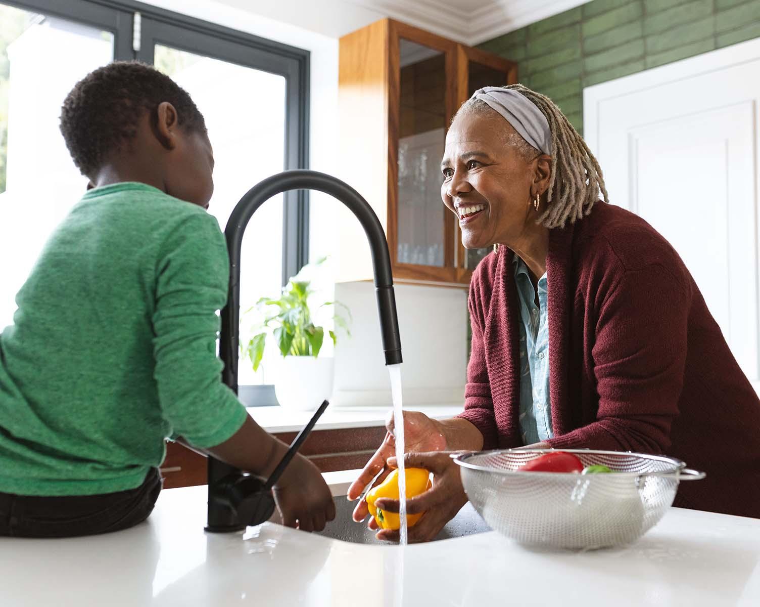 A smiling elderly woman and a boy wash vegetables in a sunny kitchen